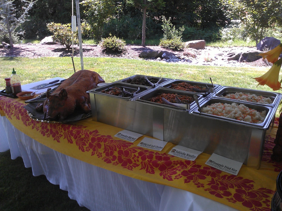 Whole roasted kalua pig on a platter next to labeled chafing trays of Hawaiian BBQ beef, loli loli chicken, yakisoba noodles, and pineapple fried rice